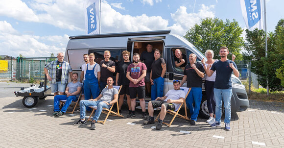 SMV Team 2023. Die Mitarbeitenden von SMV stehen an einem sonnigen Tag lachend vor einem Camper.