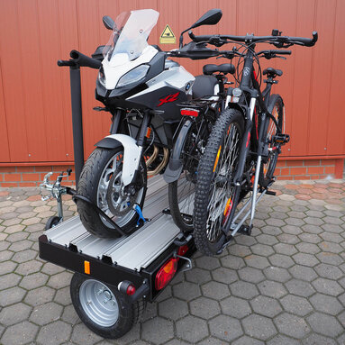 SMV Alu-Star Rolli frei stehend, schräge Seitenansicht. Maximale Beladung mit einem Motorrad auf der Plattform des Anhängers und zwei Fahrrädern auf dem optionalen Fahrradträger.