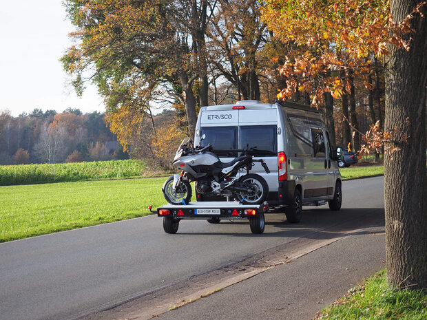 SMV Kastenwagen-Fan. Heckansicht eines Kastenwagens mit SMV-Anhänger, beladen mit einem Motorrad, fährt auf Baumallee.